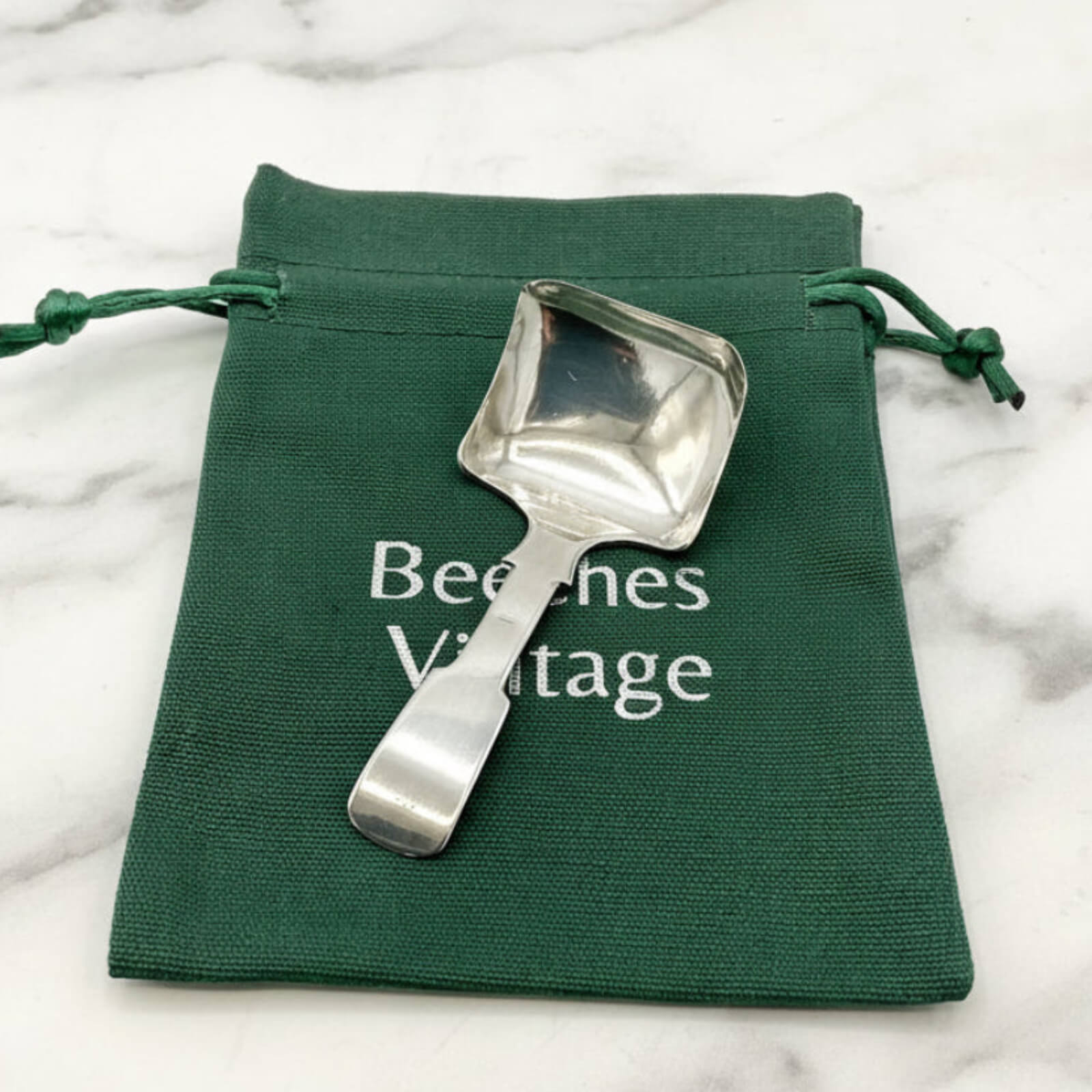 Silver scoop on a green drawstring bag with 'Beeches Vintage' text on a marble background