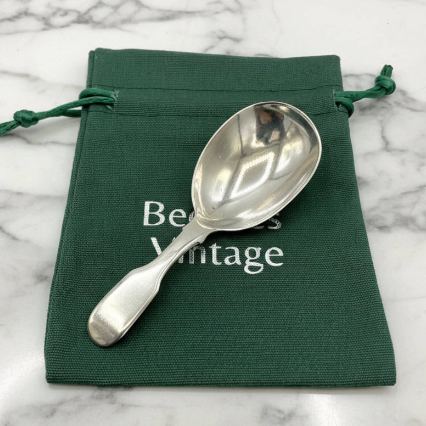 Silver spoon on a green drawstring bag with 'Beeches Vintage' text on a marble background