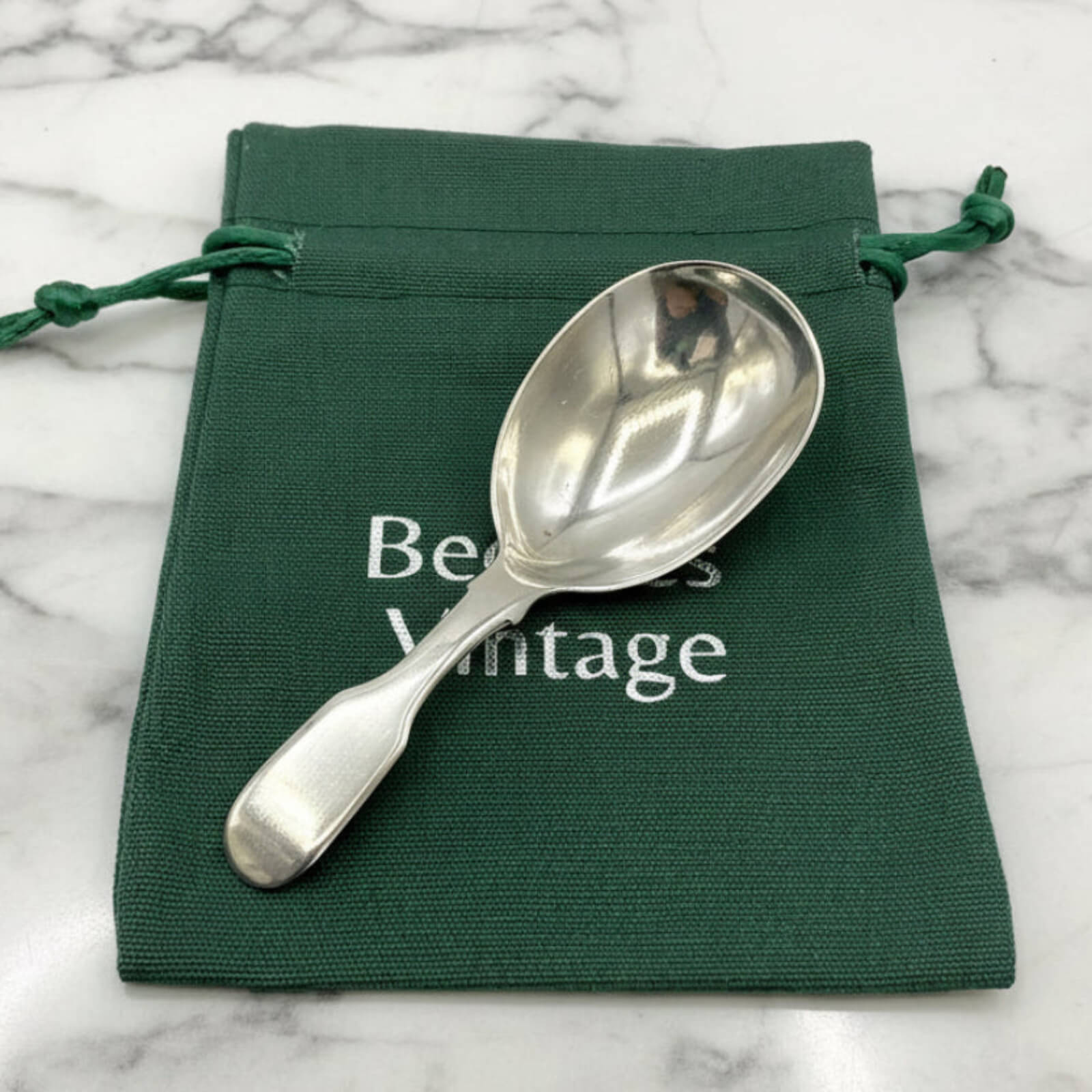 Silver spoon on a green drawstring bag with 'Beeches Vintage' text on a marble background