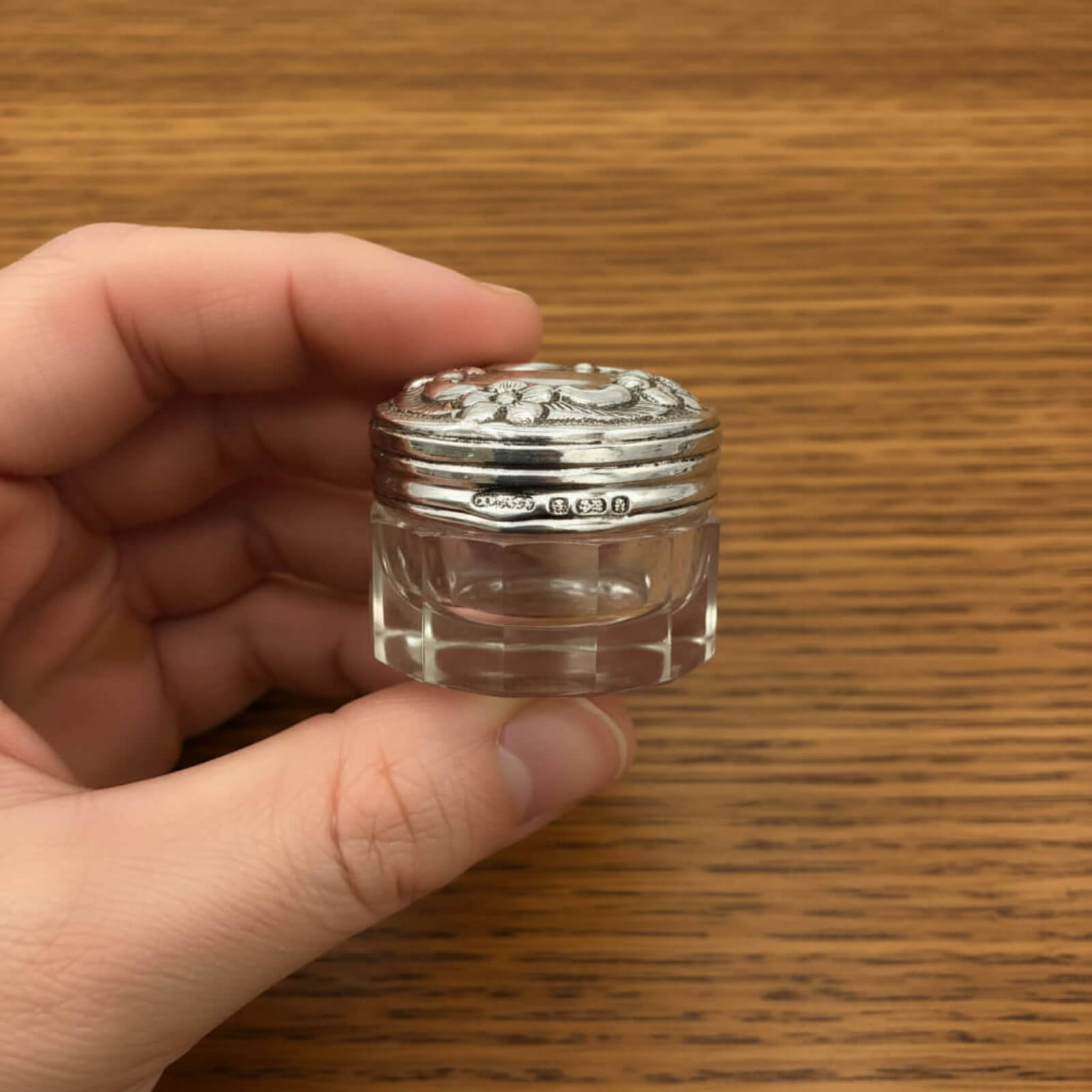Hand holding a small glass jar with a silver lid on a wooden surface