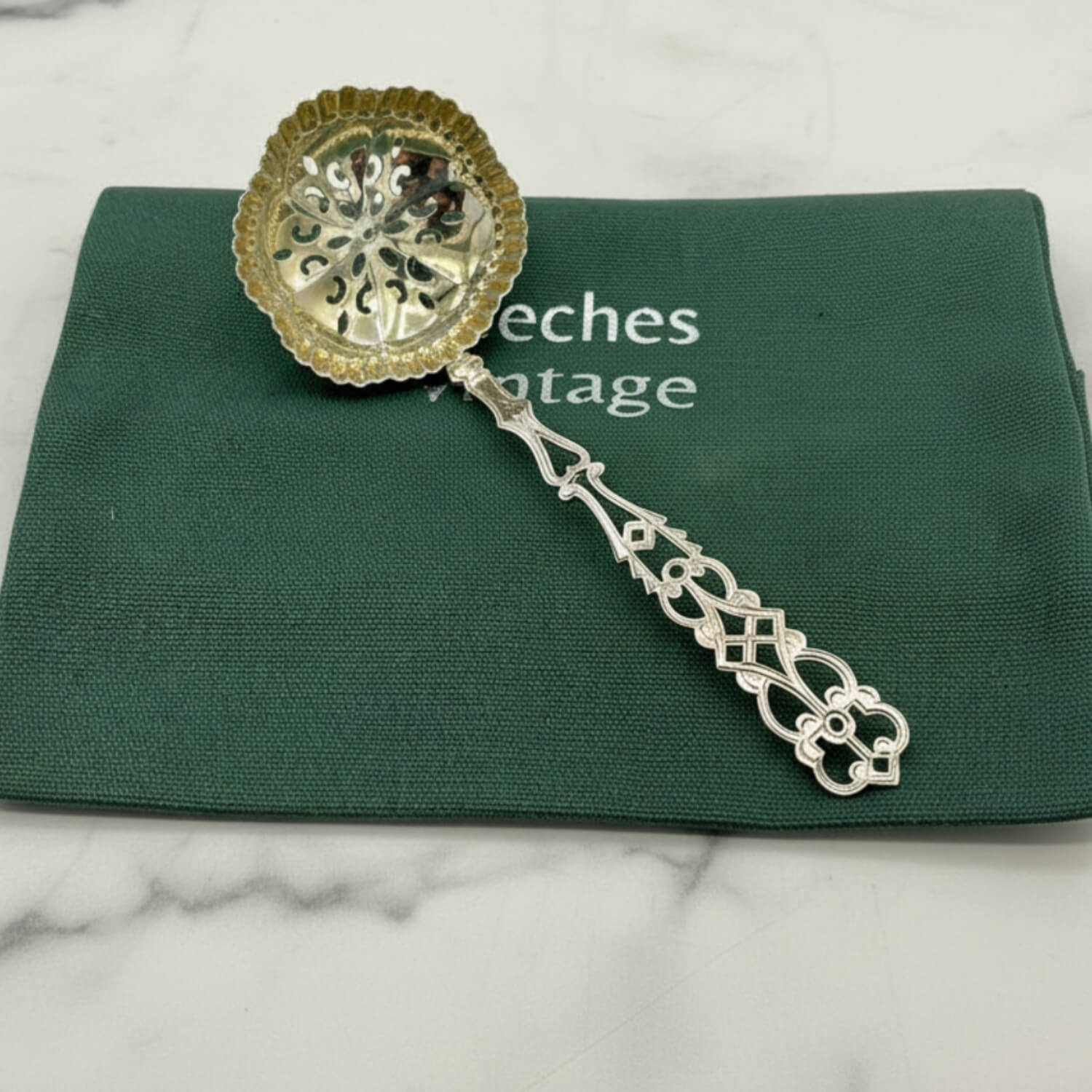 An ornate silver sugar sifter spoon with handle and a scalloped bowl, resting on a dark green velvet cloth that has the brand name 'Beeches Vintage' printed on it, set against a white marble background.