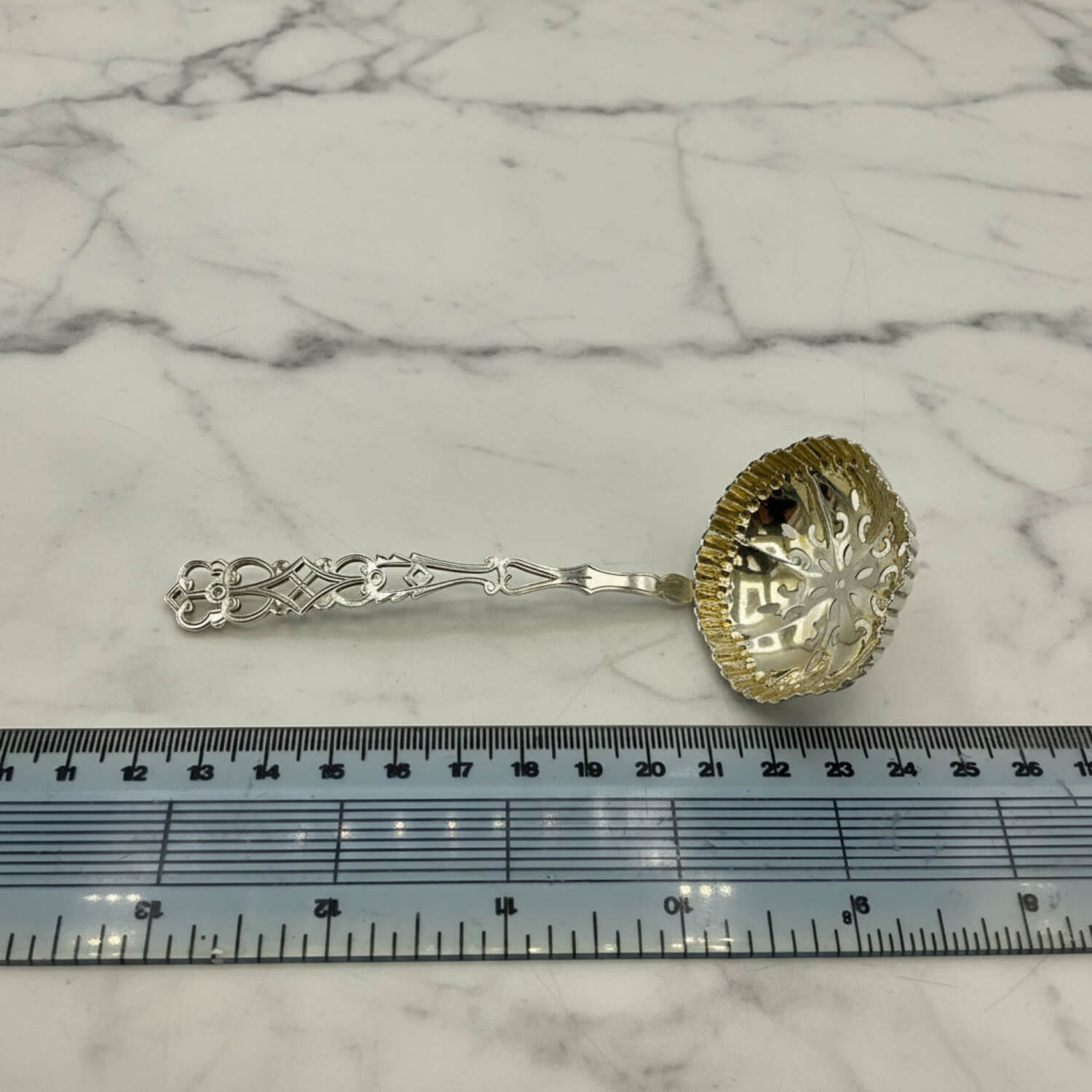 A small ornate silver sifter spoon with a decorative handle and a fluted bowl, placed on a white marble surface with a ruler underneath for scale.