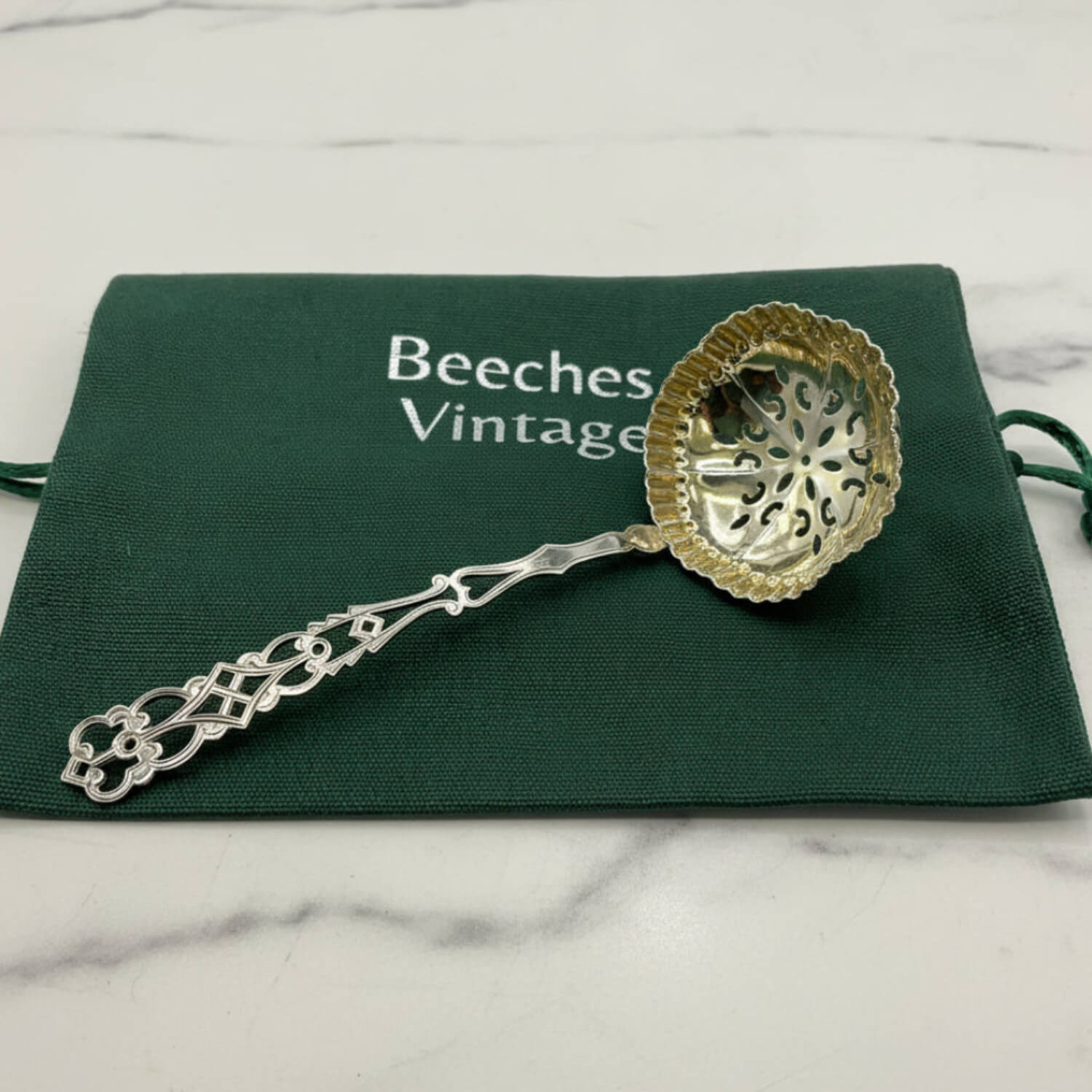 An ornate sugar sift spoon with a gold-toned perforated bowl, resting on a dark green drawstring pouch printed with 'Beeches Vintage' on a white marble surface