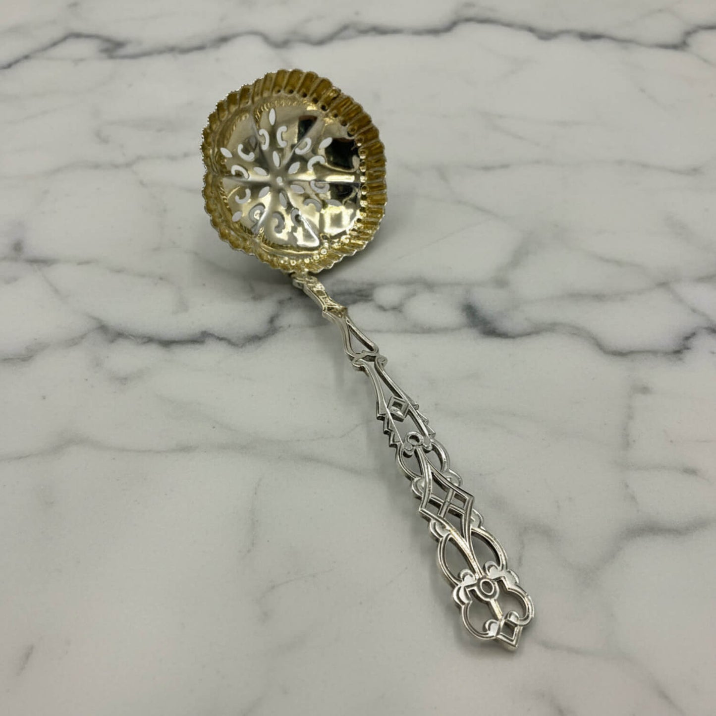 A decorative silver sifter spoon with a scalloped gold-toned bowl and an ornate silver-toned handle, placed on a white marble surface.