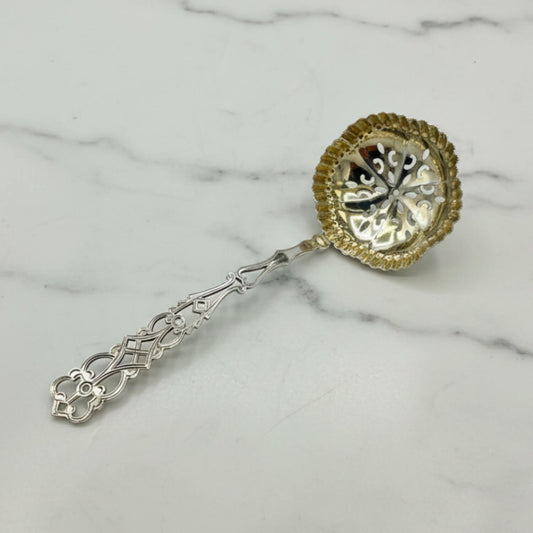 An ornate silver sugar sifter spoon with a decorative filigree handle and a gold-toned scalloped bowl, resting on a white marble surface.