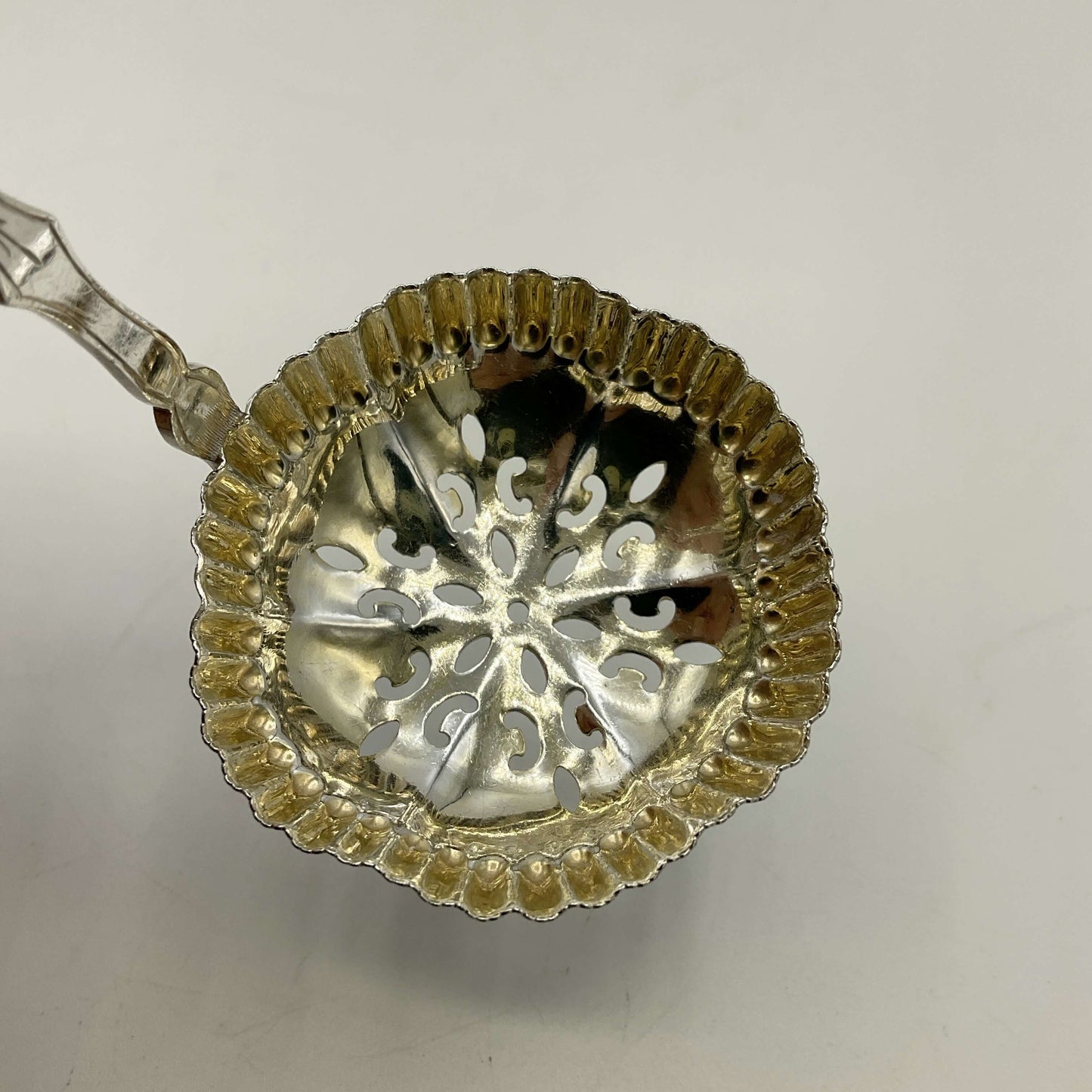 A silver sifter spoon with a scalloped rim and decorative cutout pattern, shown on a plain light background.