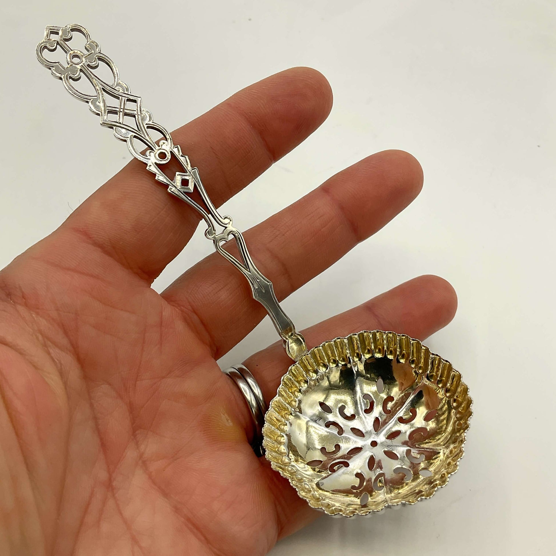 A hand holding a decorative silver sifter spoon with a gold-toned bowl, shown against a plain white background.