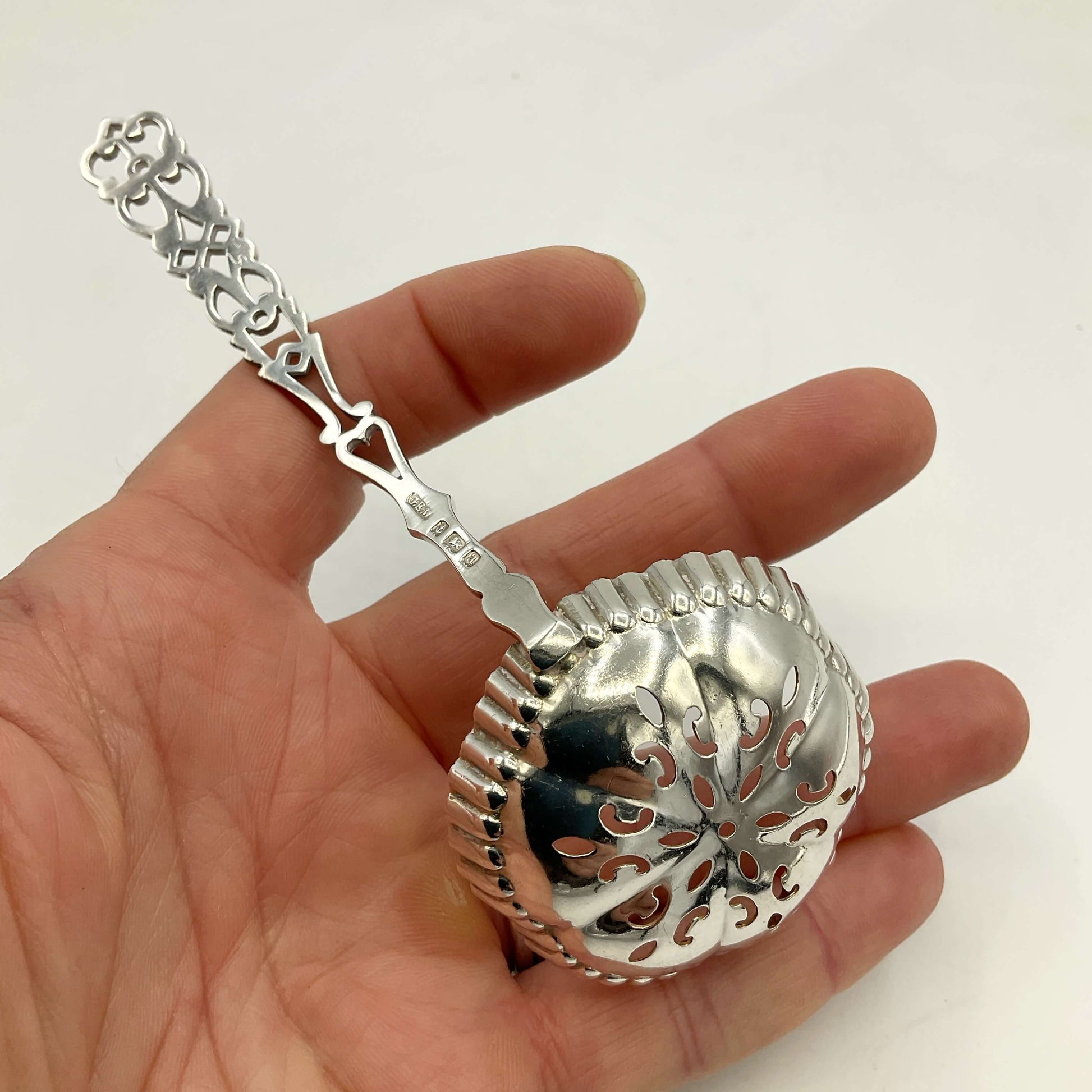 A silver sifter spoon with an ornate handle and perforated round basket, held in a person's hand against a plain white background.