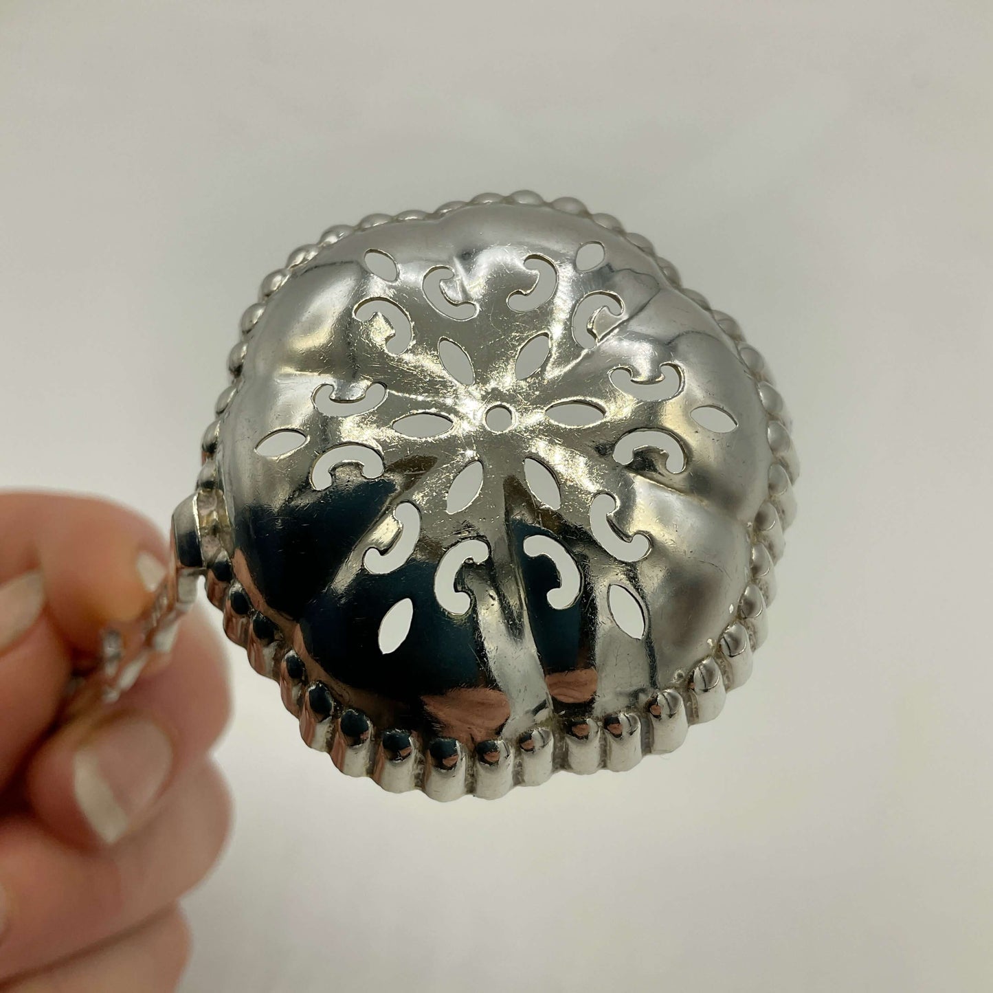 A silver sifter spoon with an ornate cutout floral pattern and beaded border, held by a hand against a plain light background.