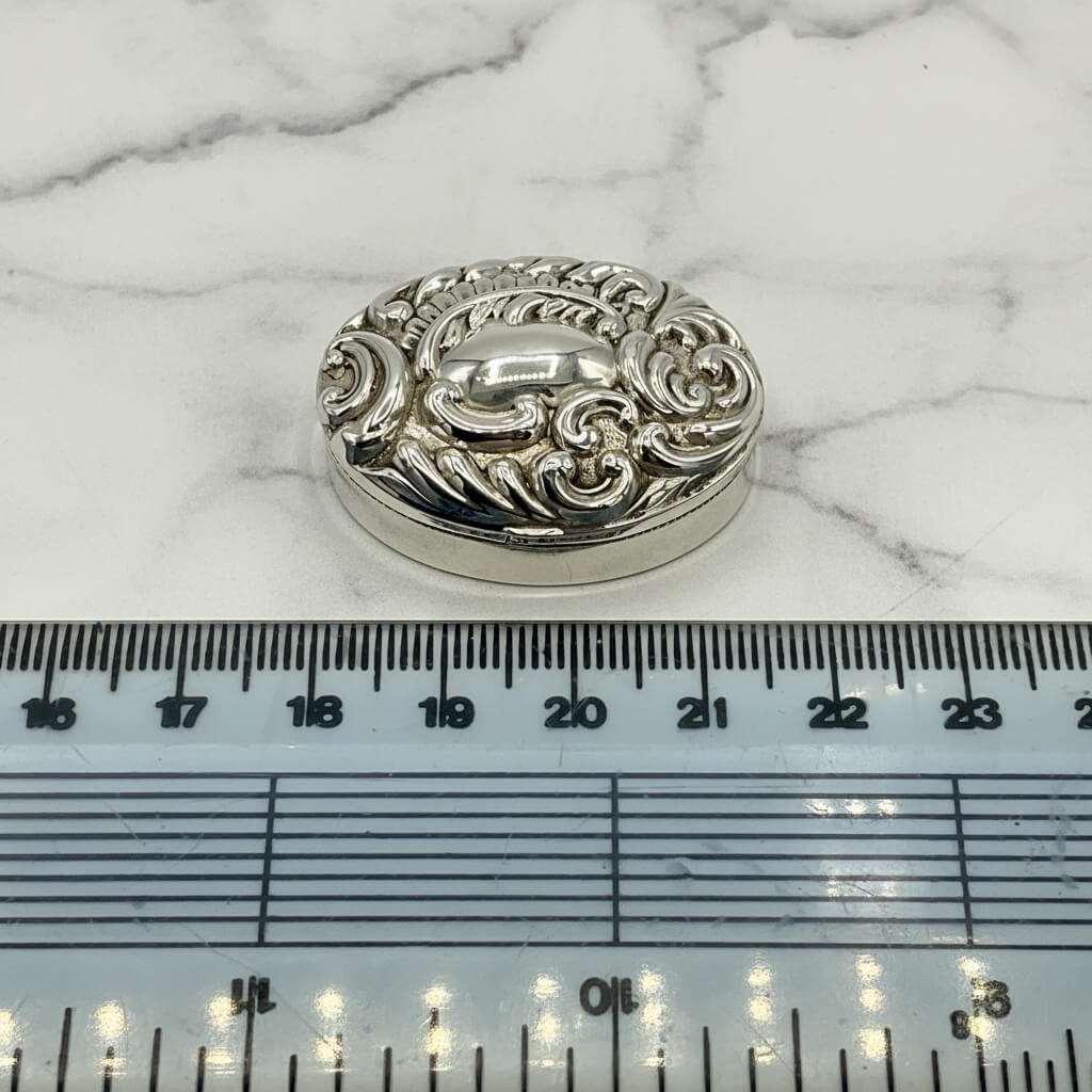 Silver decorative pill box on a marble surface with a ruler for scale.
