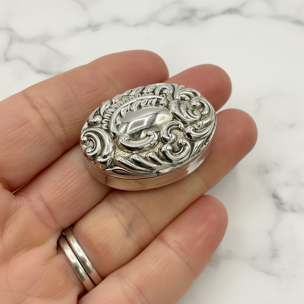 Silver decorative pill box held in a hand on a marble background