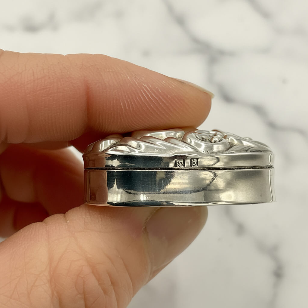 Silver decorative pill box showing hallmarks held in a hand with a blurred background
