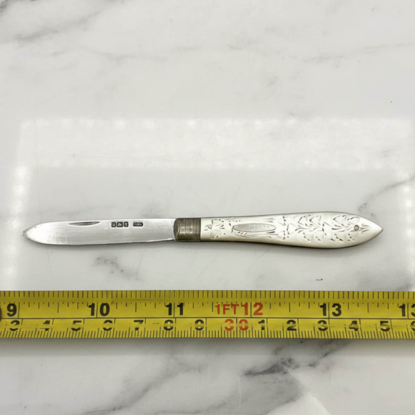 Silver fruit knife with decorative handle on a marble surface, measured by a yellow tape measure.