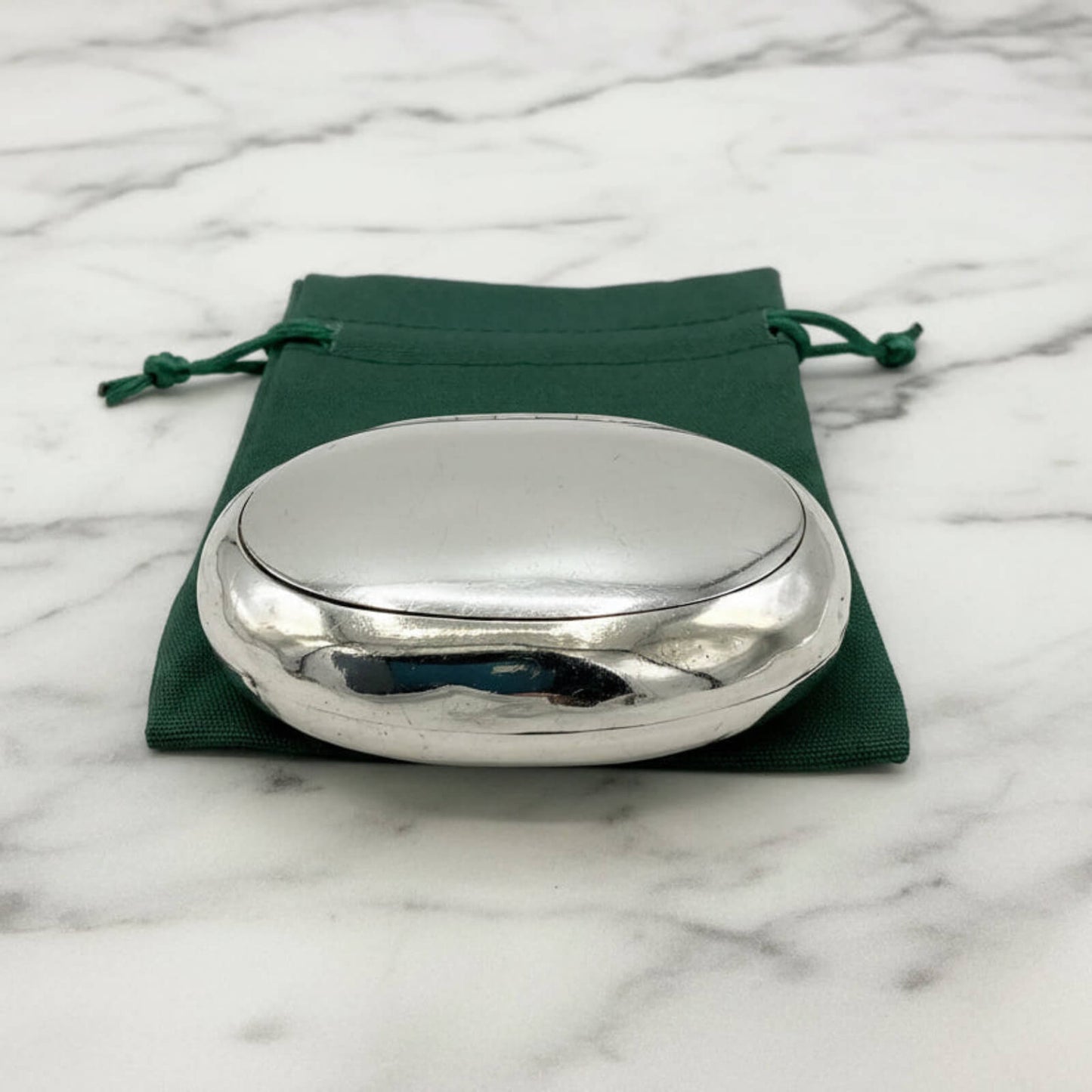 Silver oval box on a green drawstring bag with a marble background