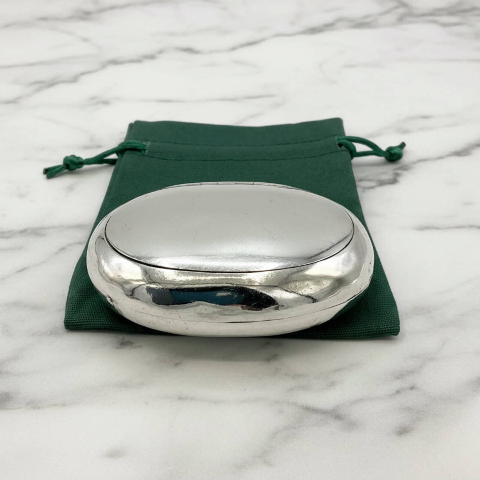 Silver oval box on a green drawstring bag with a marble background