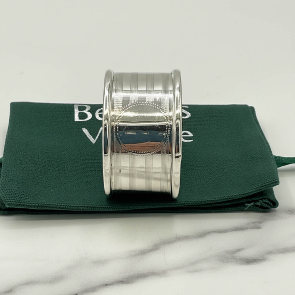 Antique 1920 Sterling Silver Napkin ring with a textured, ribbed outer band and a smooth central oval inlay, displayed on a dark green velvet pouch over a white marble surface.