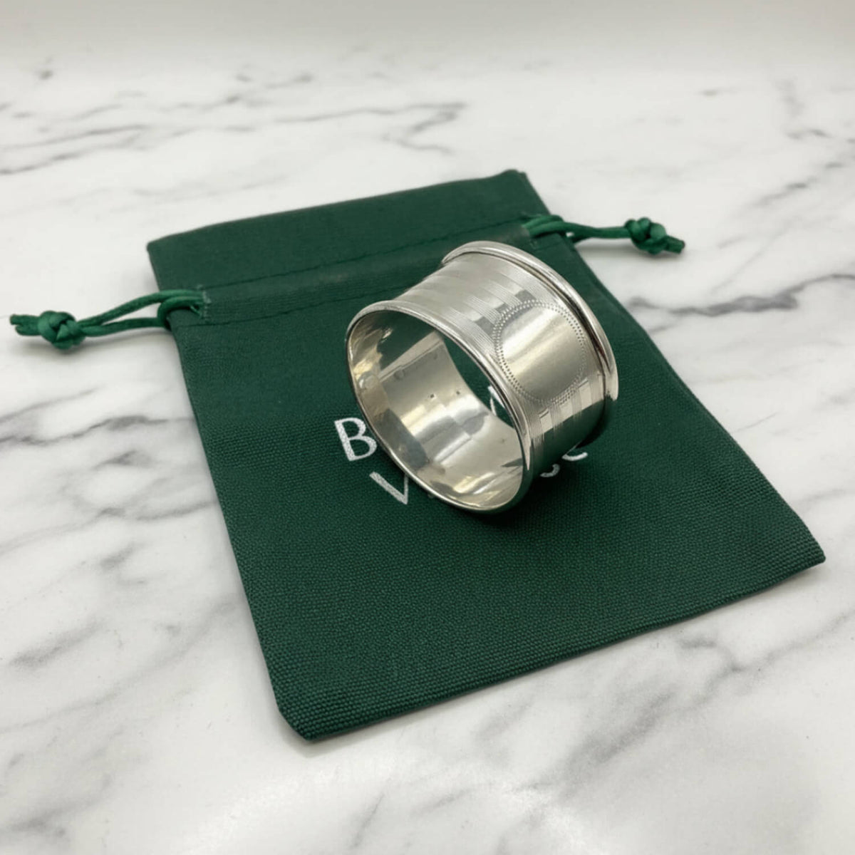 A polished Antique 1920 Sterling Silver Napkin ring with a textured outer band, resting on a dark green drawstring pouch against a white background.