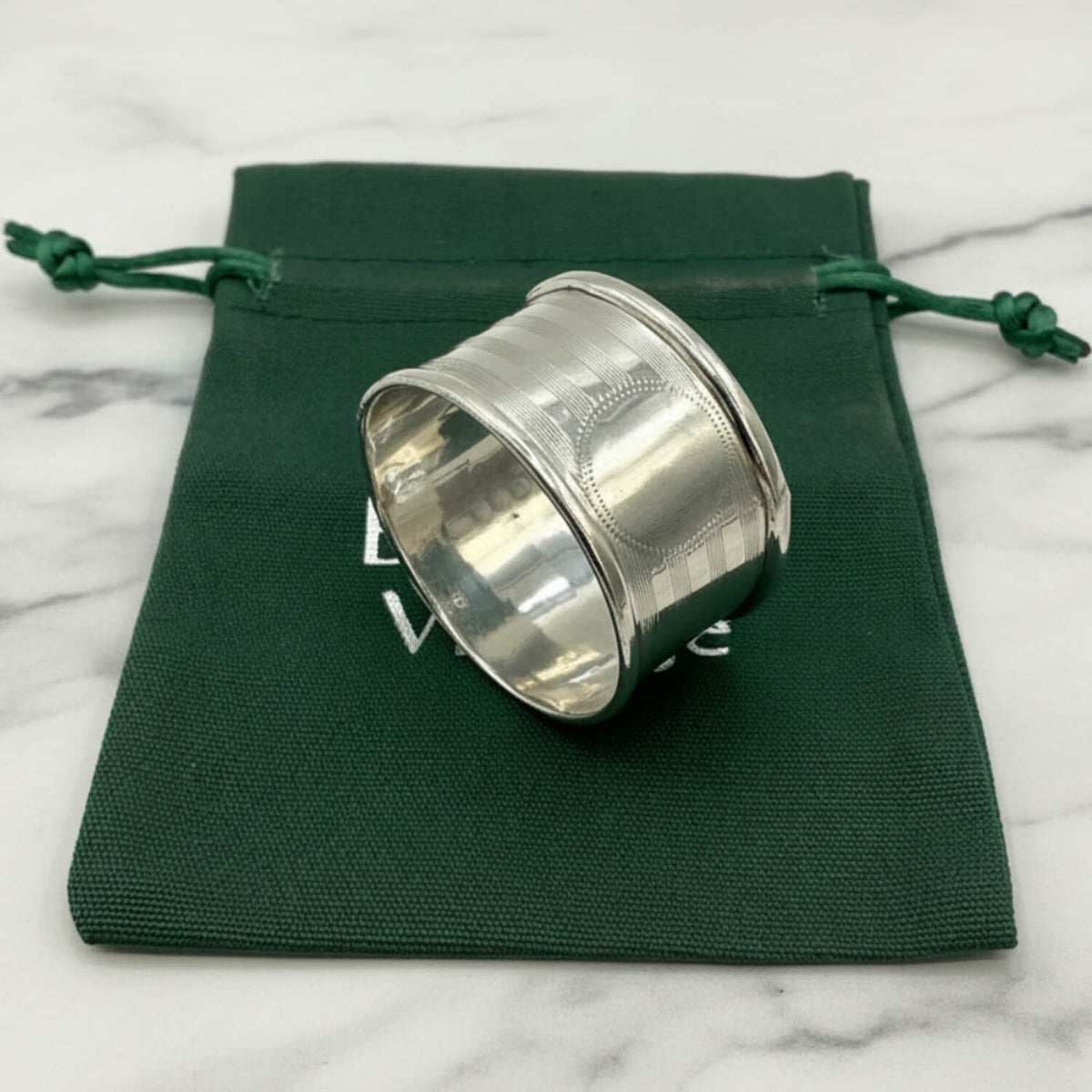 A polished Antique 1920 Sterling Silver Napkin ring with a textured outer band, resting on a dark green drawstring pouch against a white background.
