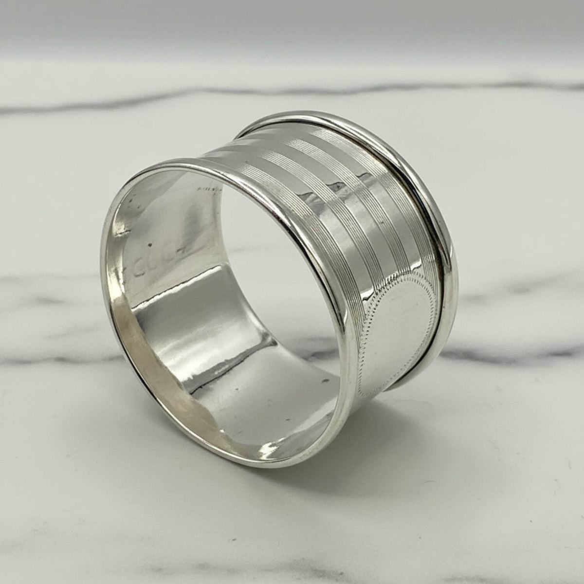 A polished Antique 1920 Sterling Silver Napkin ring with a textured, ribbed outer band and a smooth, reflective inner band, resting on a white marble surface.