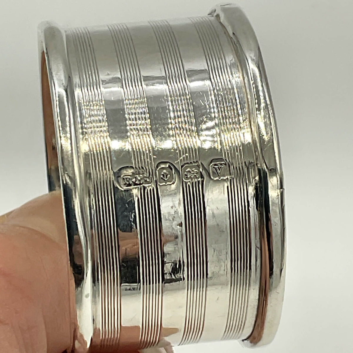 A close-up of an Antique 1920 Sterling Silver Napkin ring with engraved hallmarks, vertical lines and a polished finish, held by a hand against a plain light background.