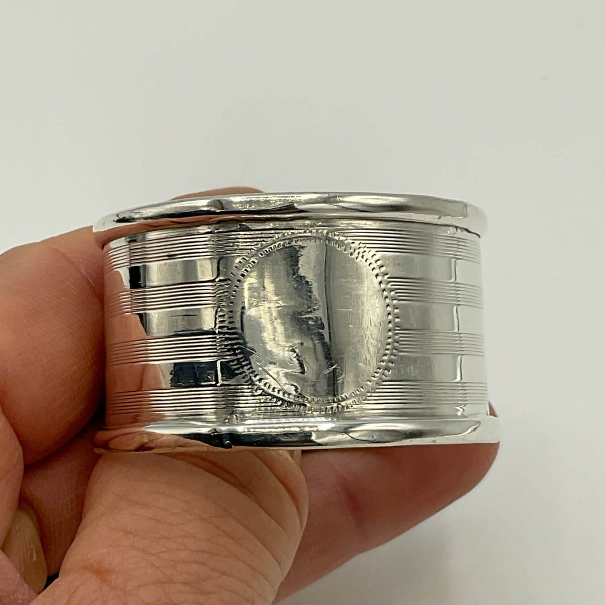 A close-up of a hand holding Antique 1920 Sterling Silver Napkin ring with engraved horizontal lines and a circular medallion on the front, against a plain light background.