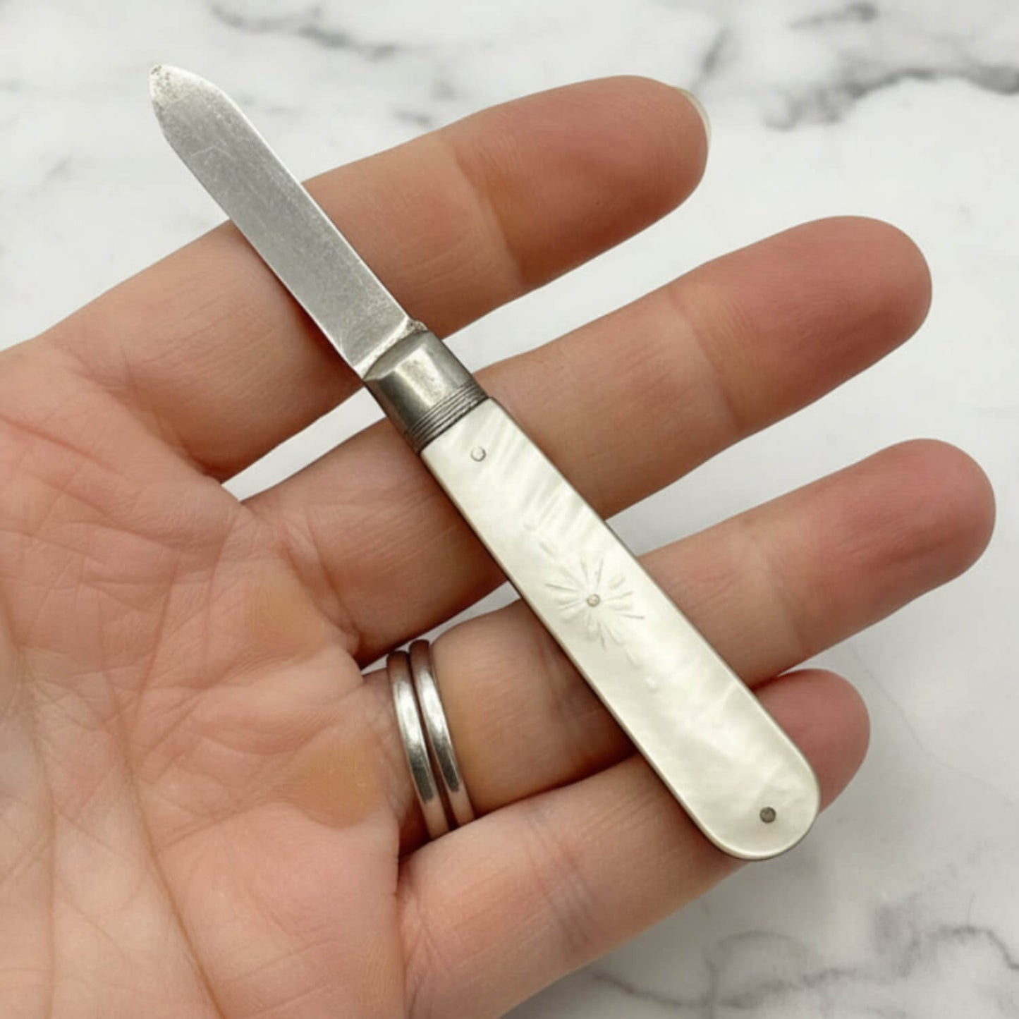 Hand holding a small silver fruit knife with an engraved handle on a marble surface.