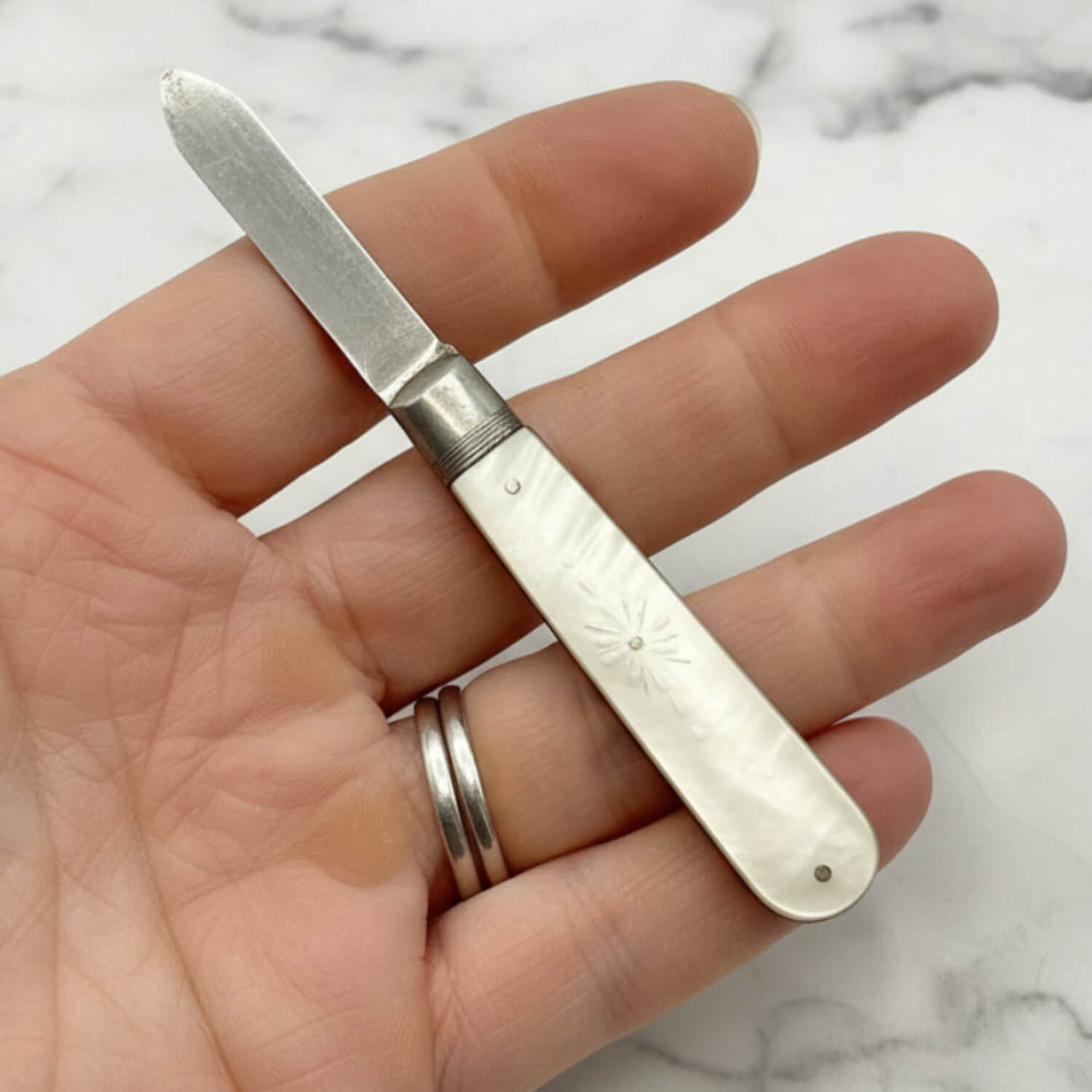 Hand holding a small silver fruit knife with an engraved handle on a marble surface.