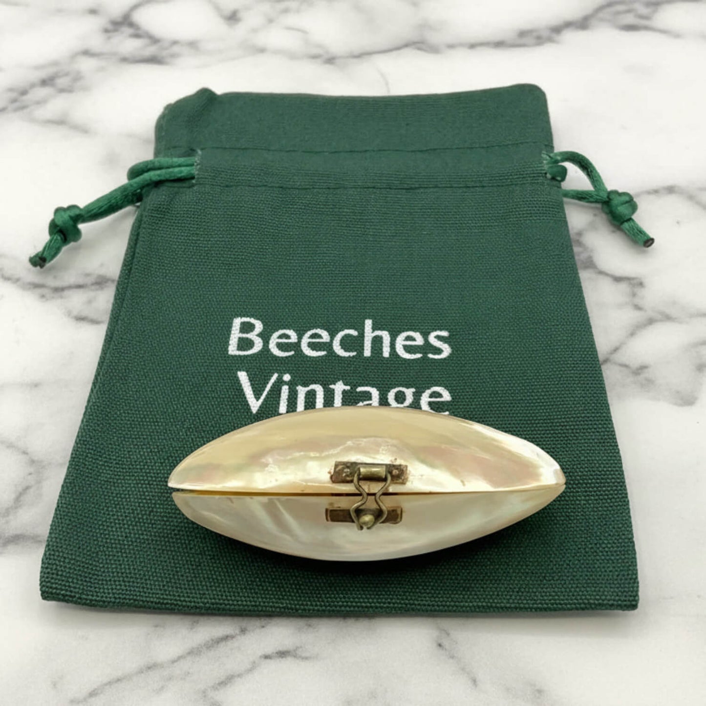 Shell-shaped pill box on a green drawstring bag with 'Beeches Vintage' text on a marble background