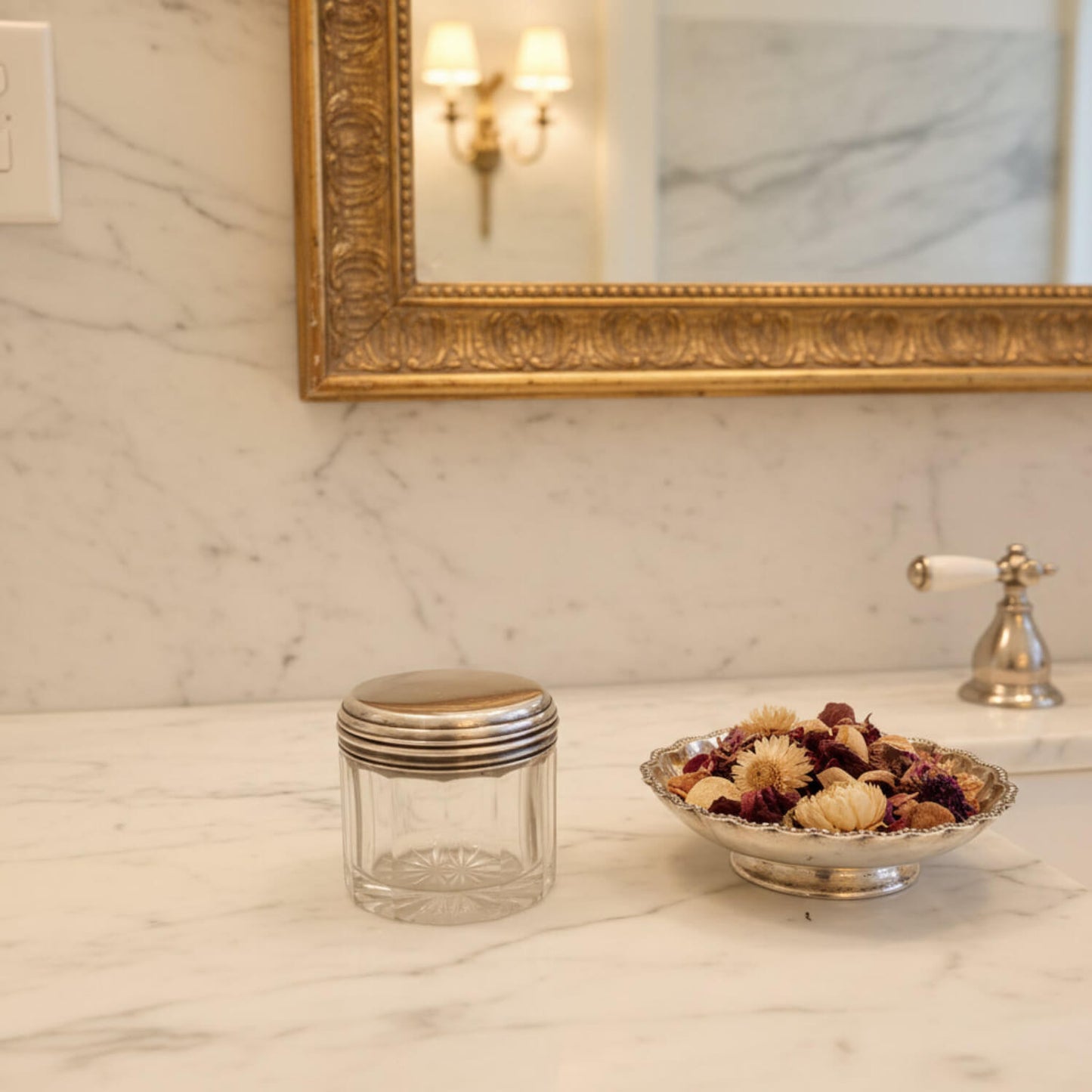 Glass jar with silver lid and decorative bowl of flowers on a marble countertop with a gold-framed mirror in the background.