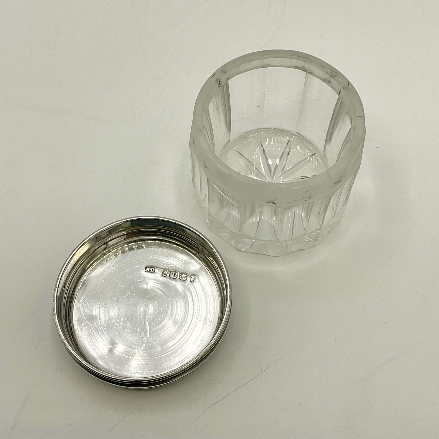 Clear glass jar with silver lid upside down showing hallmarks on a light grey background