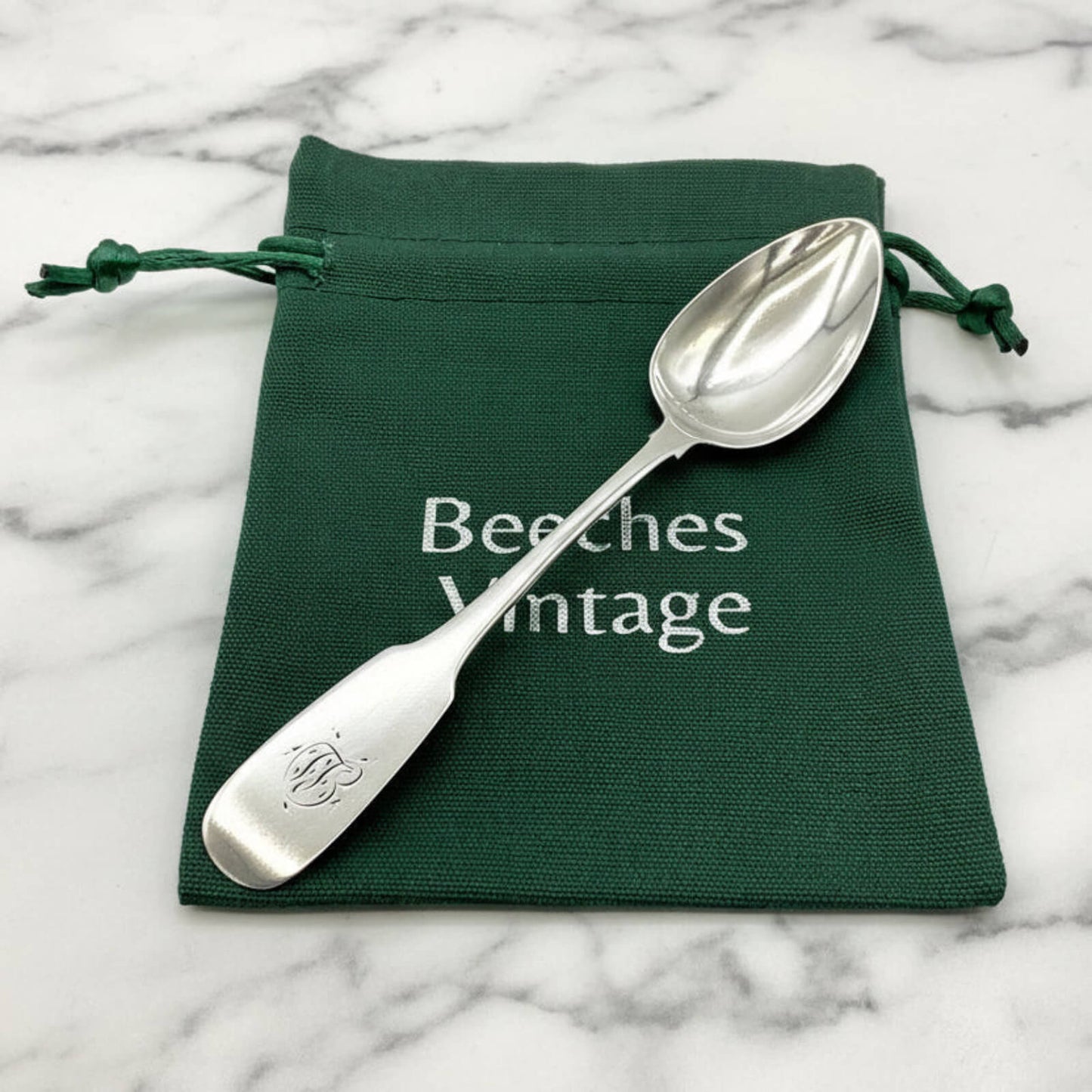 A silver spoon on a green drawstring bag with 'Beeches Vintage' branding on a marble surface.