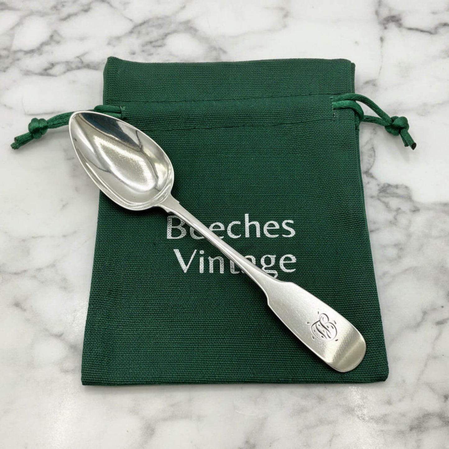 Silver spoon on a green drawstring bag with 'Beeches Vintage' branding on a marble surface.
