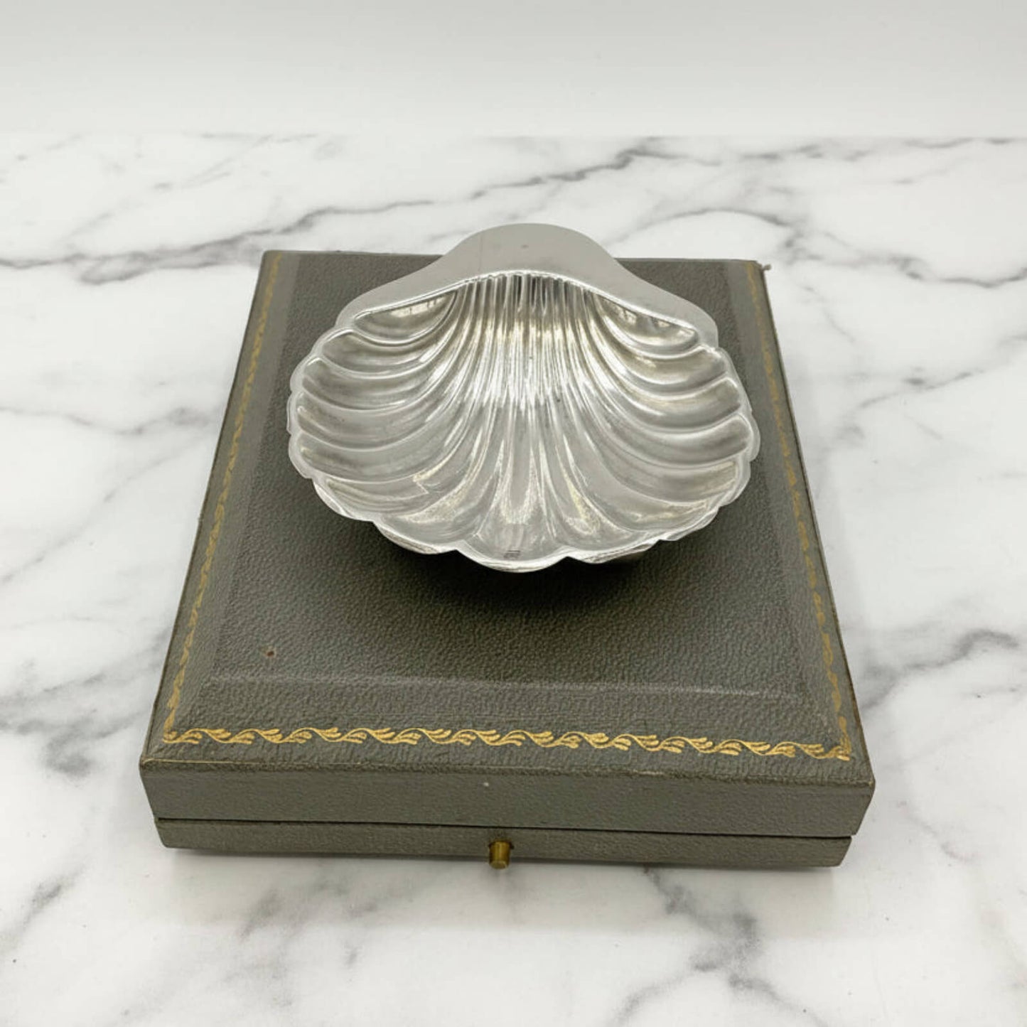 Silver shell-shaped dish on a gray box with gold trim against a marble background