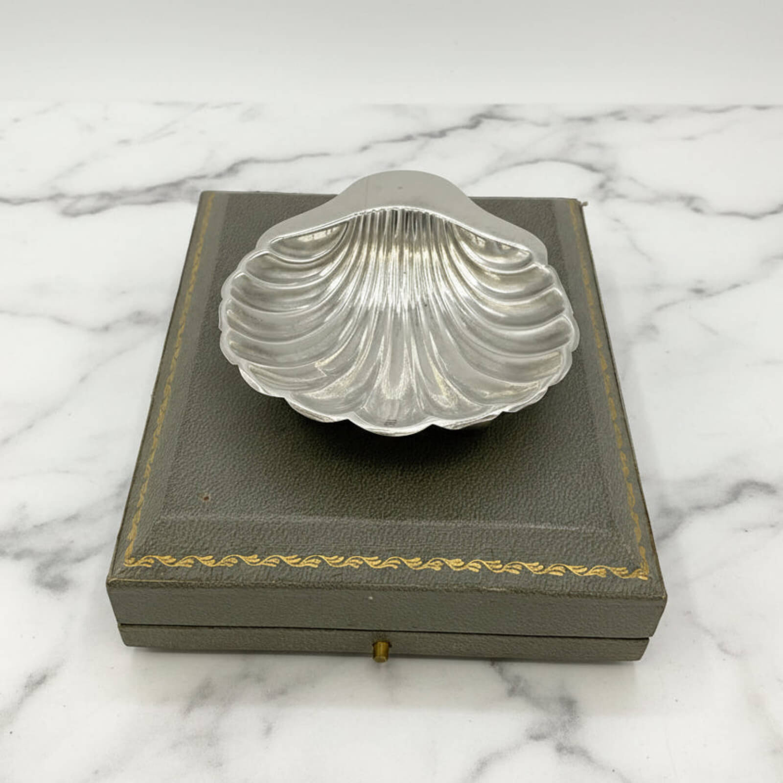 Silver shell-shaped dish on a gray box with gold trim against a marble background