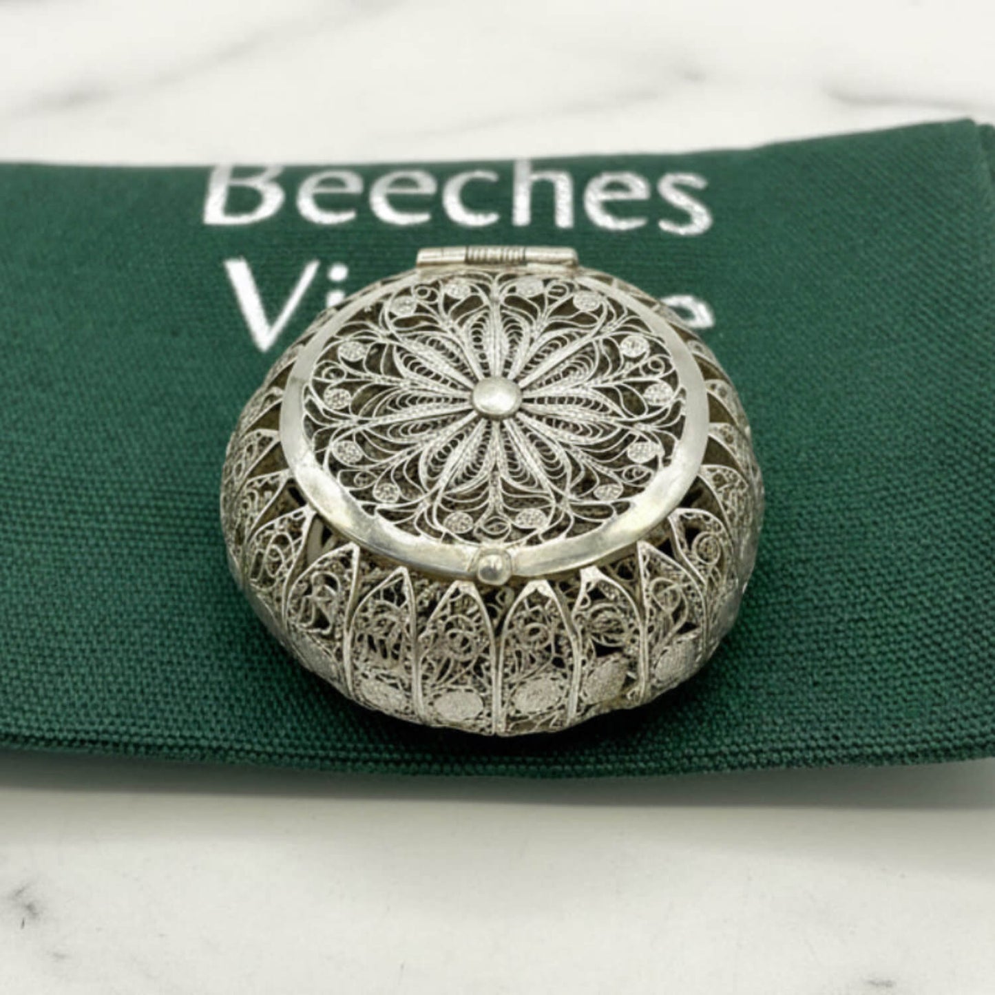 Silver filigree compact mirror on a green fabric background with 'Beeches' branding.
