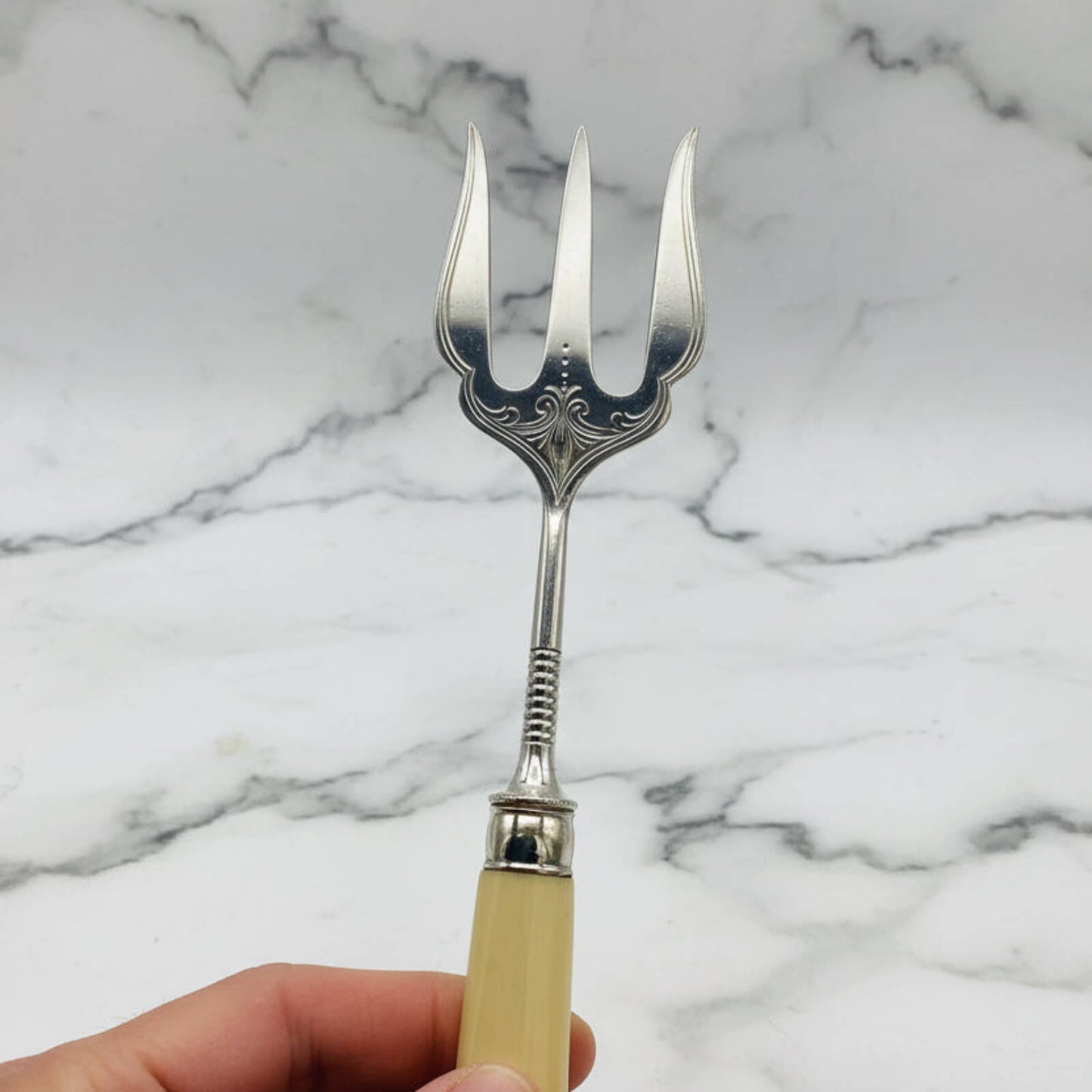 Silver decorative fork with a beige handle held against a marble background
