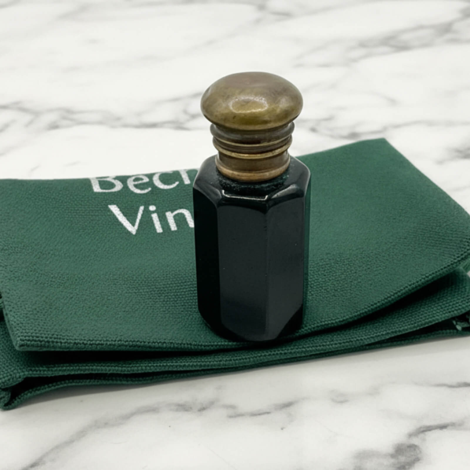 Black ink bottle with a brass cap on green fabric with 'Beeches Vintage' branding, on a marble surface