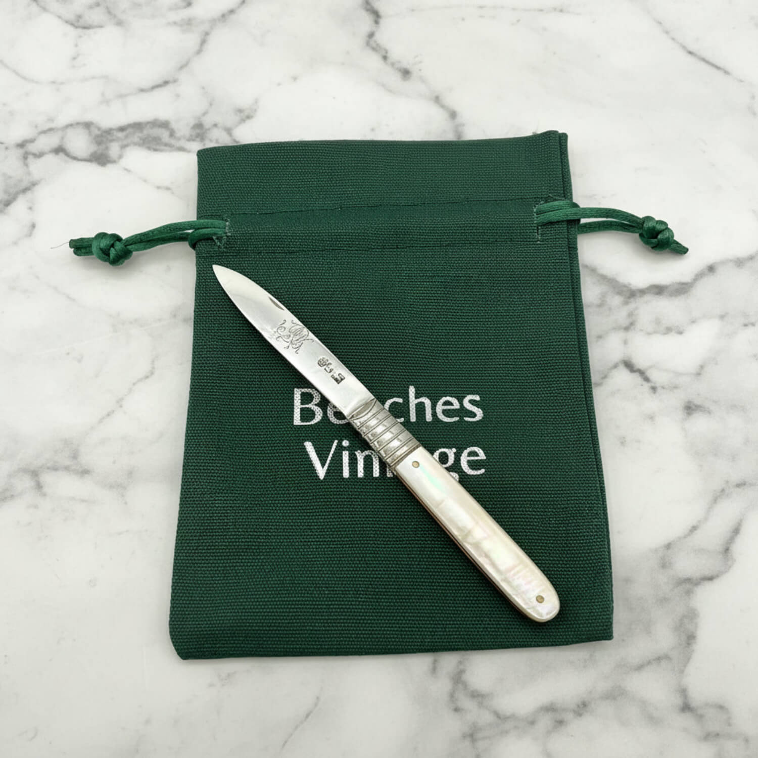 A silver fruit knife with an mother-of-pearl handle rests diagonally on a dark green drawstring pouch. The pouch features the white printed text 'Beeches Vintage'.