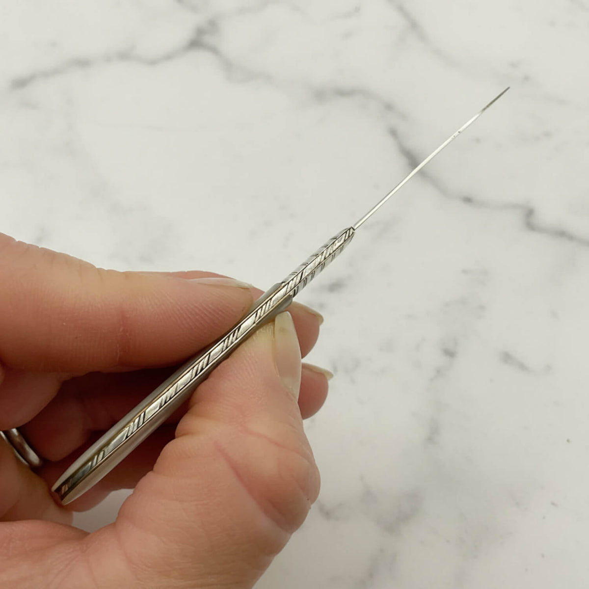 A close-up of a hand holding a long, slender silver fruit knife with a textured spine handle against a white marble background.