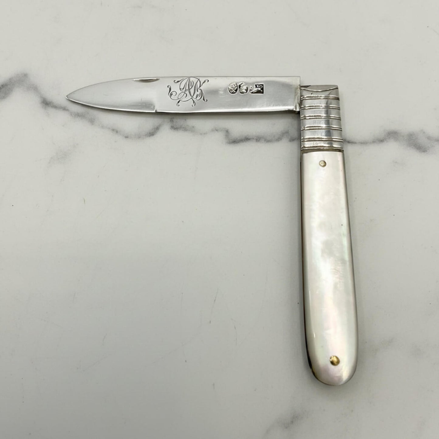 Antique  silver fruit knife with a silver blade and mother of pearl handle, featuring engraved initials and a ribbed metal bolster, displayed on a light gray background.