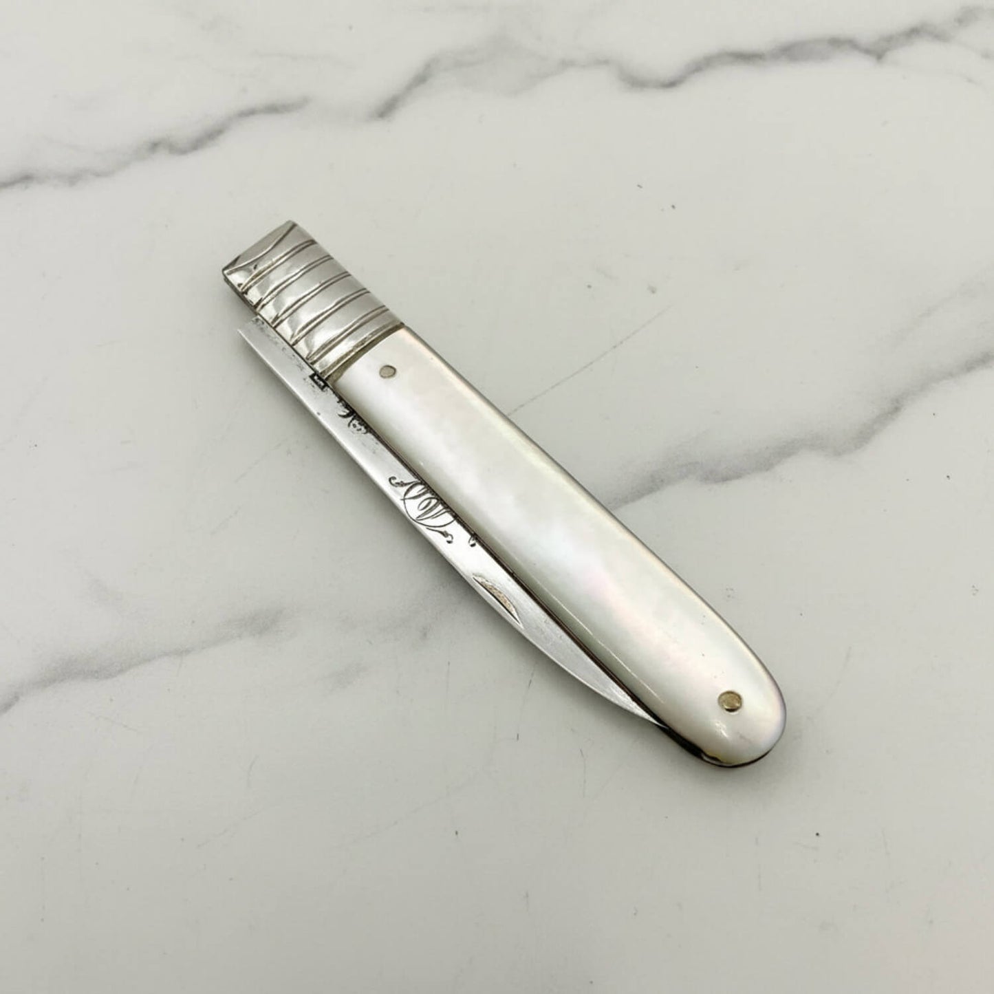 Antique silver knife with a mother-of-pearl handle and a silver collar, resting diagonally on a white marble surface.
