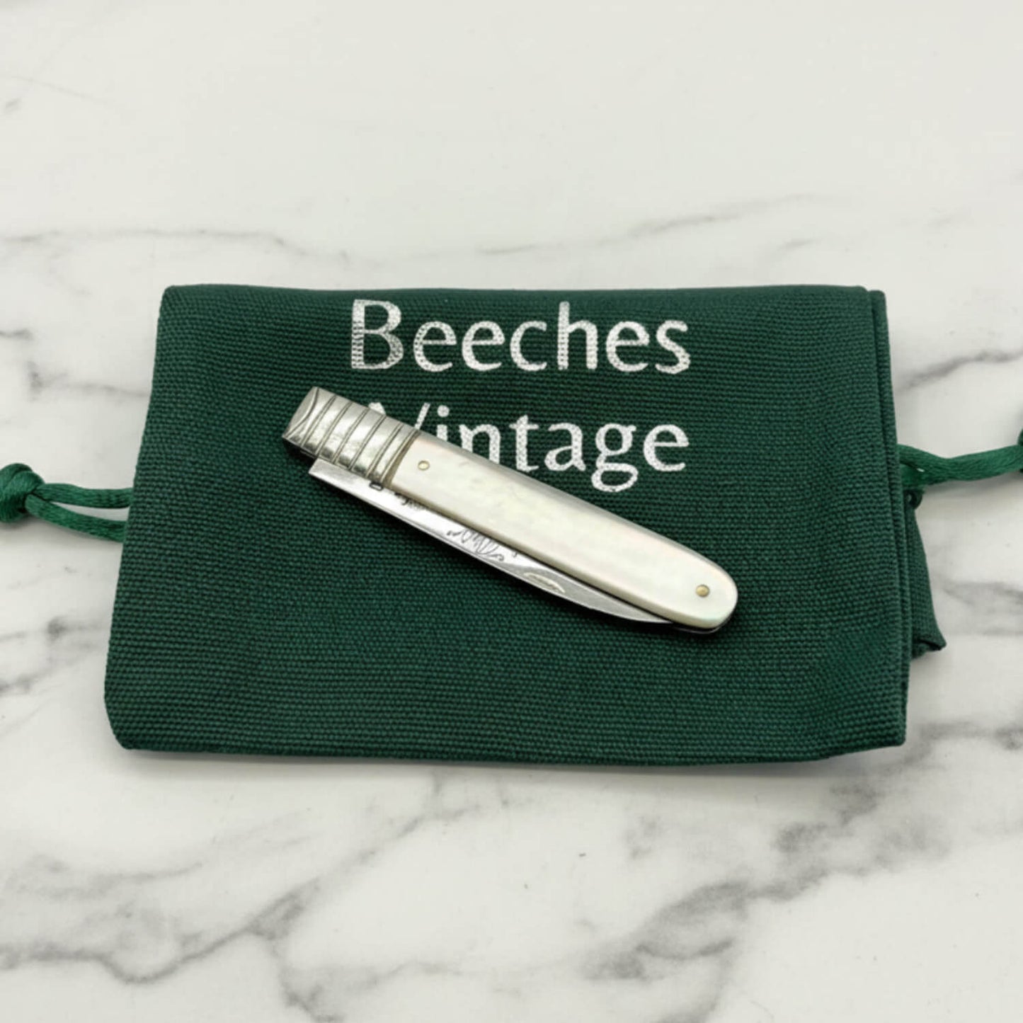 Antique silver fruit knife with a white mother-of-pearl handle, resting on a dark green canvas drawstring pouch with 'Beeches Vintage' printed in silver.