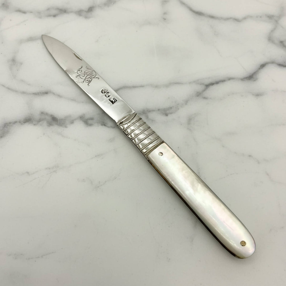 An silver fruit knife with a silver blade and handle, featuring a mother-of-pearl inlay and a textured grip, shown on a neutral light gray background.