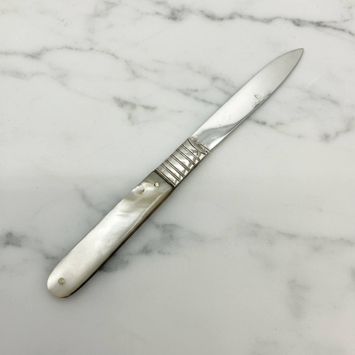 An antique silver fruit knife with a mother-of-pearl handle, displayed diagonally on a white and gray marble surface.