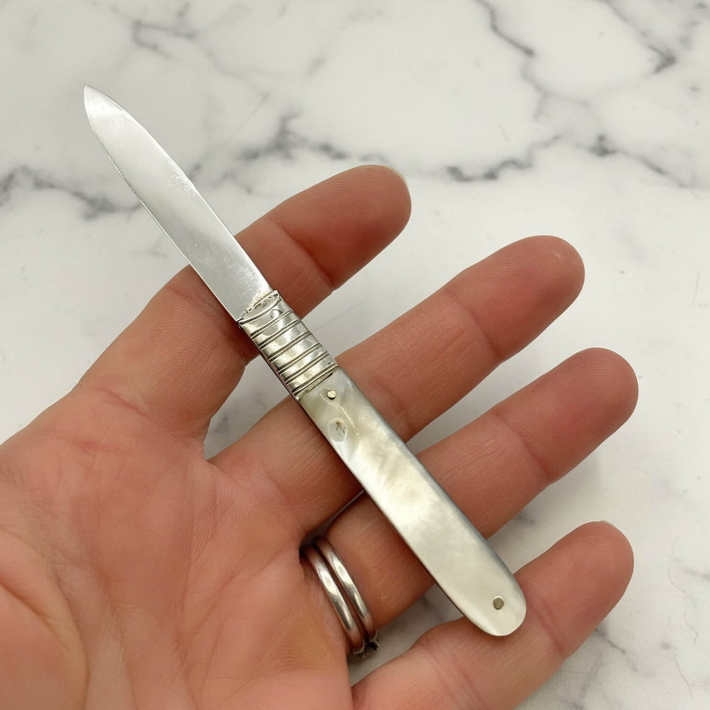A silver fruit knife with a mother of pearl handle, held in a person's hand against a white marble background.