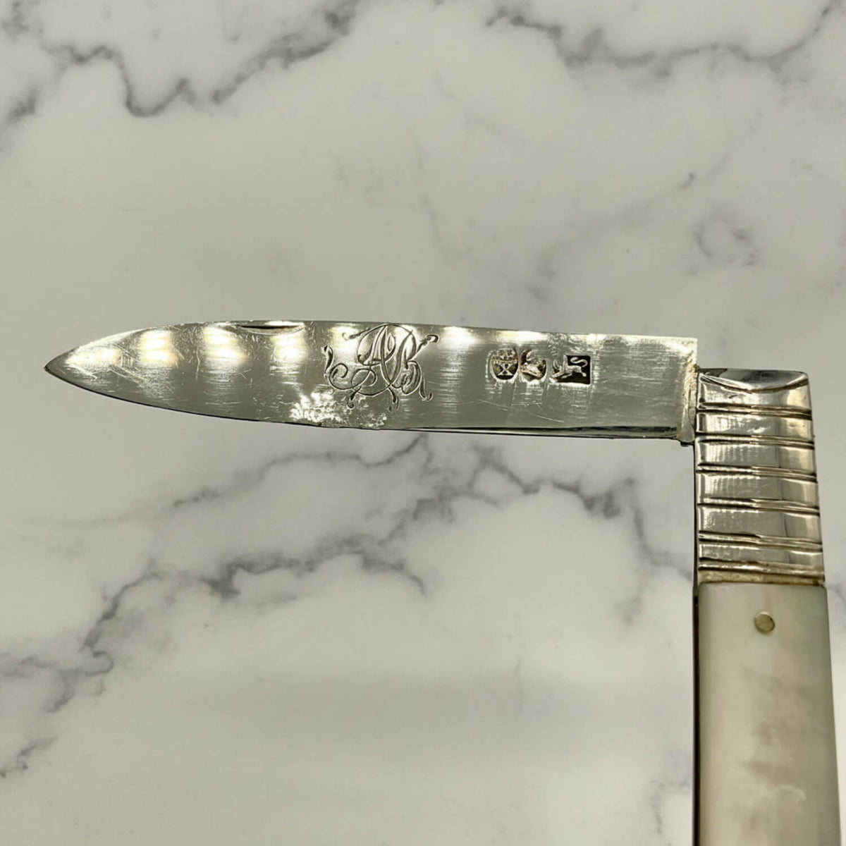 Antique silver knife with engraved initials and sterling silver hallmarks mark, and a mother-of-pearl handle, shown against a neutral light gray background.