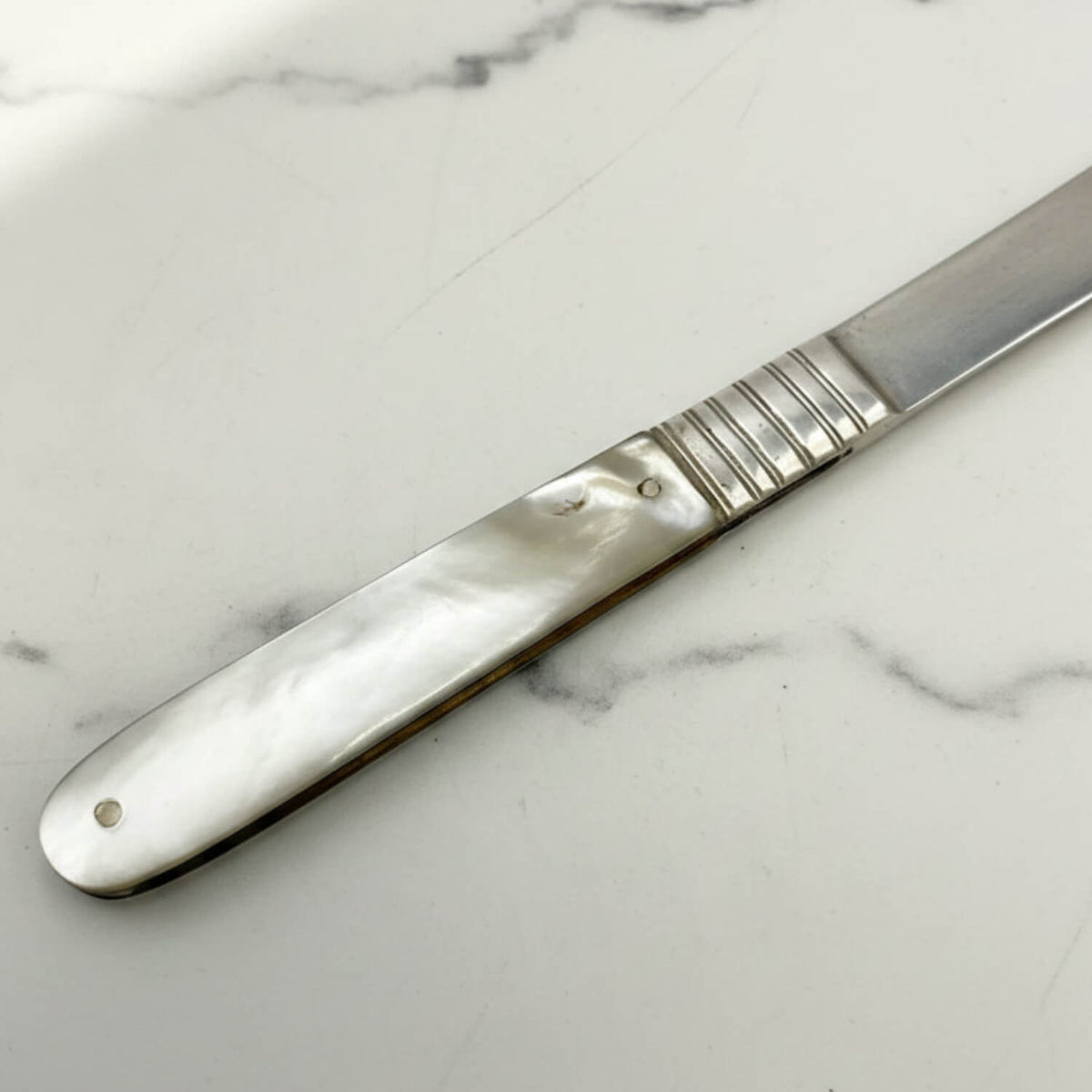 A silver fruit knife with a textured handle and a mother-of-pearl inlay, resting diagonally on a white marble surface.