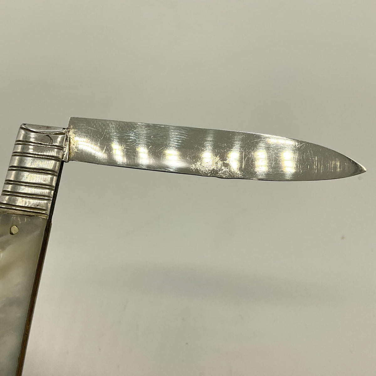 A close-up of a silver knife blade and part of a textured silver and mother of pearl handle against a plain light gray background.