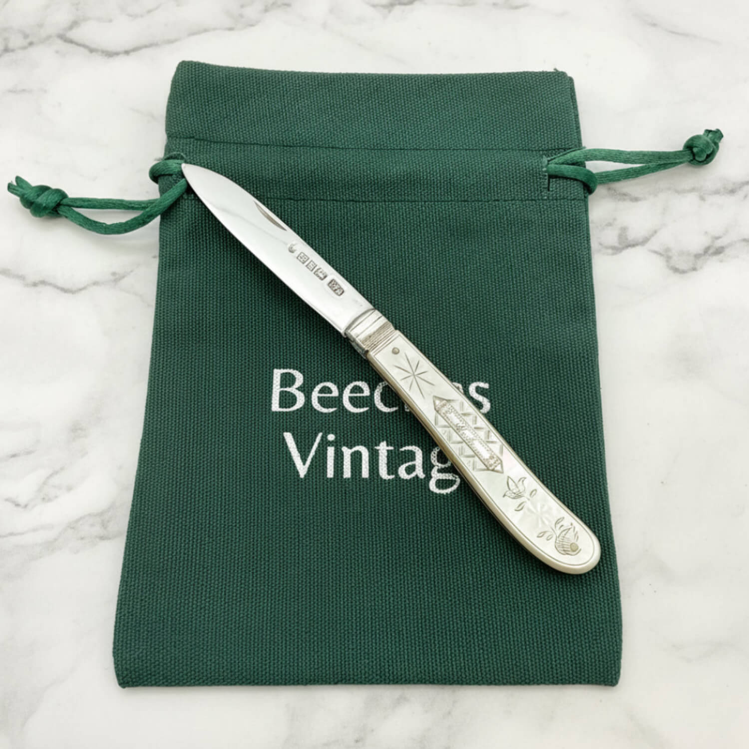 An antique silver knife with an engraved handle rests diagonally on a dark green drawstring pouch. The pouch features the text 'Beeches Vintage' printed in white.