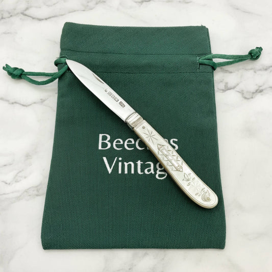 An antique silver knife with an engraved handle rests diagonally on a dark green drawstring pouch. The pouch features the text 'Beeches Vintage' printed in white.